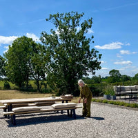Person standing next to outdoor furniture on a gravel area with trees and blue sky in the background, arranging rustic grainsack bench cushions