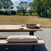 Outdoor setting with a wooden table, wicker basket, and rustic grainsack bench cushion on a gravel surface with trees and blue sky in the background.