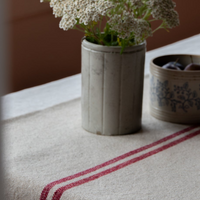 A rustic French red stripe grainsack table runner laid out on a table with a vase containing flowers on top and a small bowl with fruit in the background, on a linen tablecloth.