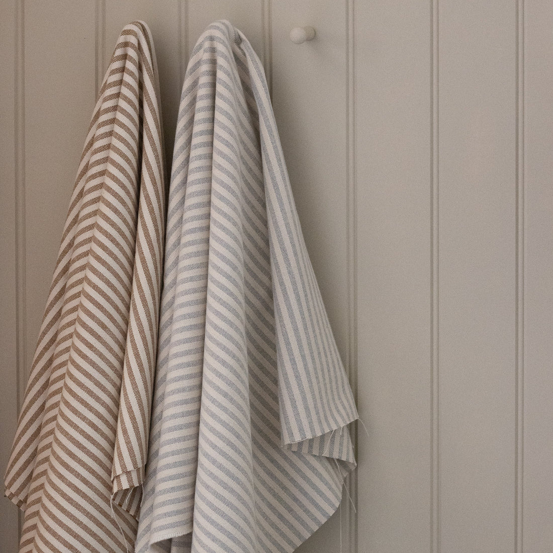 Two ticking striped fabric lengths hanging on a hook against a beige paneled wall.