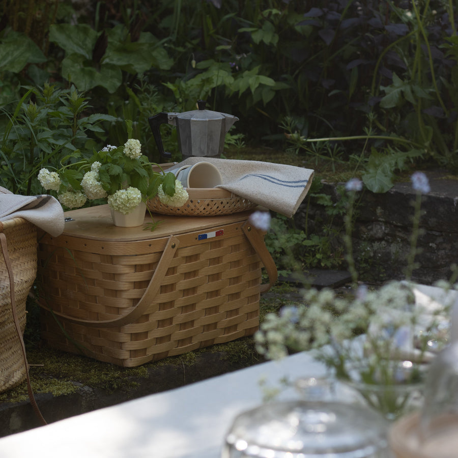Outdoor setting with a picnic basket, flowers, and a glass container on a table.