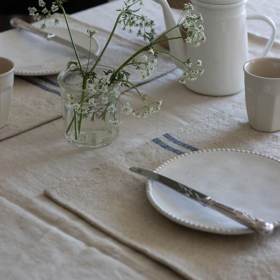 Table setting with white teapot, cup, and glass vase with flowers on a beige tablecloth.