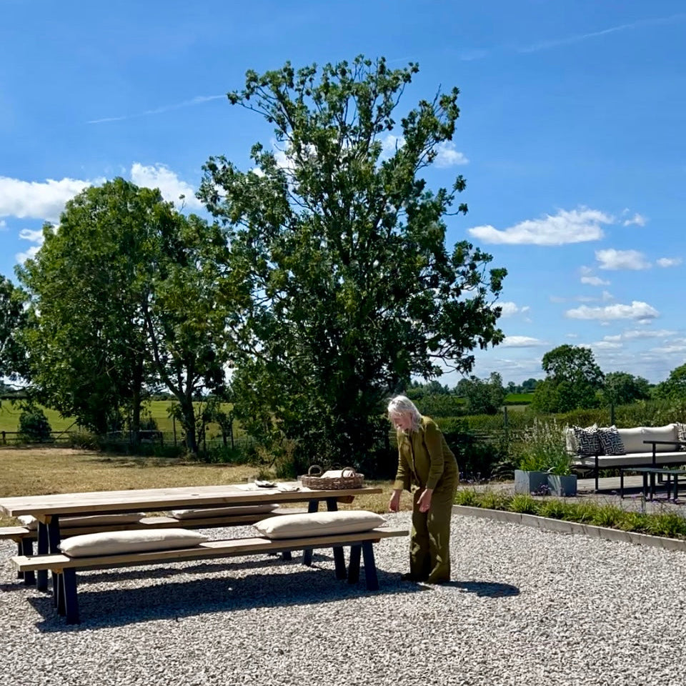 Person standing next to outdoor furniture on a gravel area with trees and blue sky in the background, arranging rustic grainsack bench cushions