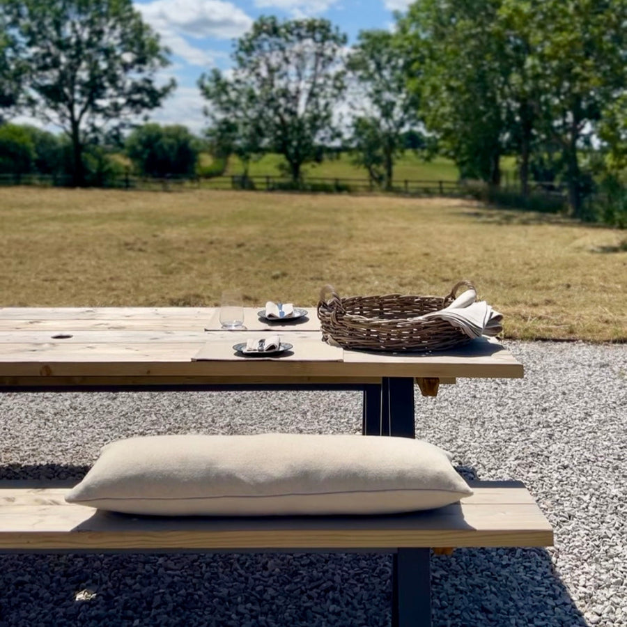 Outdoor setting with a wooden table, wicker basket, and  rustic grainsack bench cushion on a gravel surface with trees and blue sky in the background.