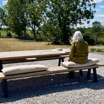 Person sitting on a bench in an outdoor setting with trees and open space featuring rustic bench cushions